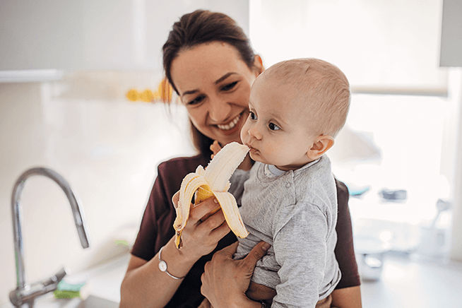 Mamá cargando a un bebé y lo alimenta con una banana en el espacio de la cocina.