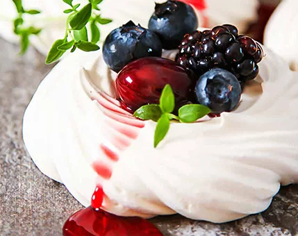 A close-up of a delicate meringue dessert topped with fresh blueberries, blackberries, and cherries. The setting features a rustic surface with vibrant red syrup dripping onto the base. Green herb sprigs add a fresh touch to the composition, enhancing the visual appeal.