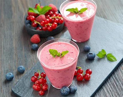 Two glasses of vibrant pink berry smoothies are served on a slate tray, garnished with fresh mint leaves. Surrounding the glasses are bowls and scattered berries, including blueberries, raspberries, and red currants, creating a colorful and appetizing display. The setting features a wooden table, adding a rustic touch to the presentation.