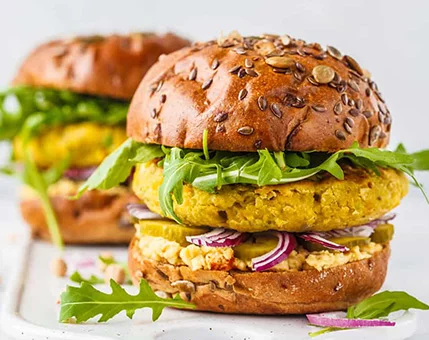 A close-up of a vegetarian burger featuring a golden chickpea patty, fresh arugula, and sliced red onions. The seeded bun adds texture and visual appeal, while hummus and pickles enhance the flavor profile. The setting is bright and clean, emphasizing the fresh ingredients.