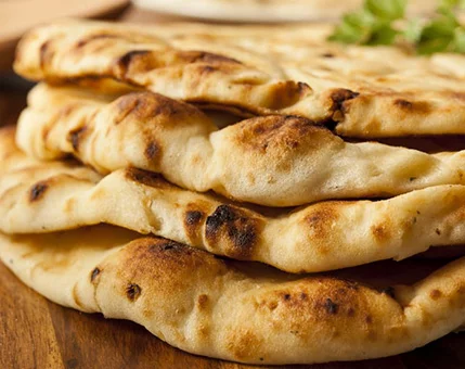 A close-up view of a stack of golden-brown naan bread, showcasing its soft texture and charred spots. The bread is placed on a wooden surface, with fresh herbs visible in the background. The image highlights the artisanal quality and rustic appeal of the baked goods.