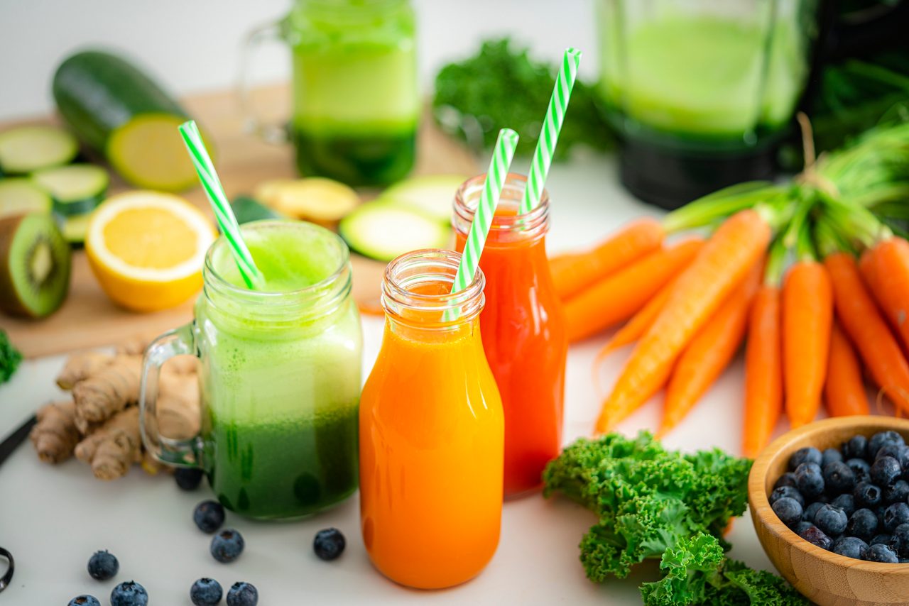 Drei Säfte aus Karotte, Orange und grünem Gemüse stehen mit Strohhalmen neben frischem Obst und Gemüse auf einem Tisch.