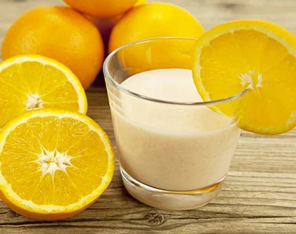 A glass of creamy orange smoothie is garnished with a fresh orange slice. Surrounding the glass are whole and halved oranges placed on a rustic wooden surface. The vibrant orange color and natural texture create a refreshing and healthy visual appeal.