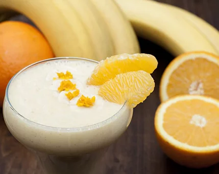 A creamy smoothie served in a glass, garnished with orange slices and zest. The setting includes fresh bananas and oranges on a wooden surface, emphasizing a natural and healthy vibe. The vibrant colors of yellow and orange create a refreshing and inviting mood.