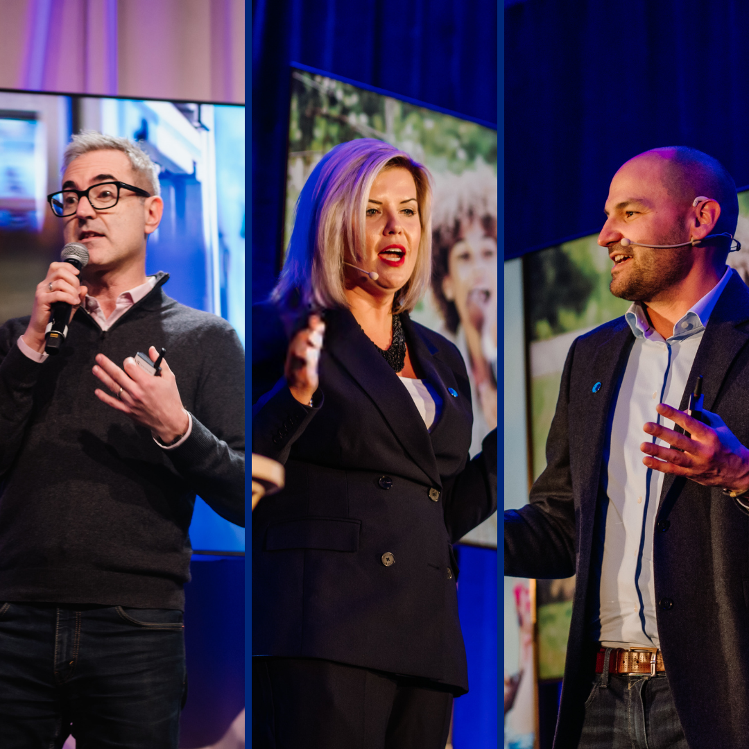 Three Danone Canada leaders stand on stage speaking at an event. From left to right: a man in a black blazer with short, grey straight hair (Frederic Guichard), a woman black blazer and blonde hair (Natalie Holloway), and a man with black blazer and a shaved head (Iannick Melancon). They're all wearing a microphone close to their mouth.