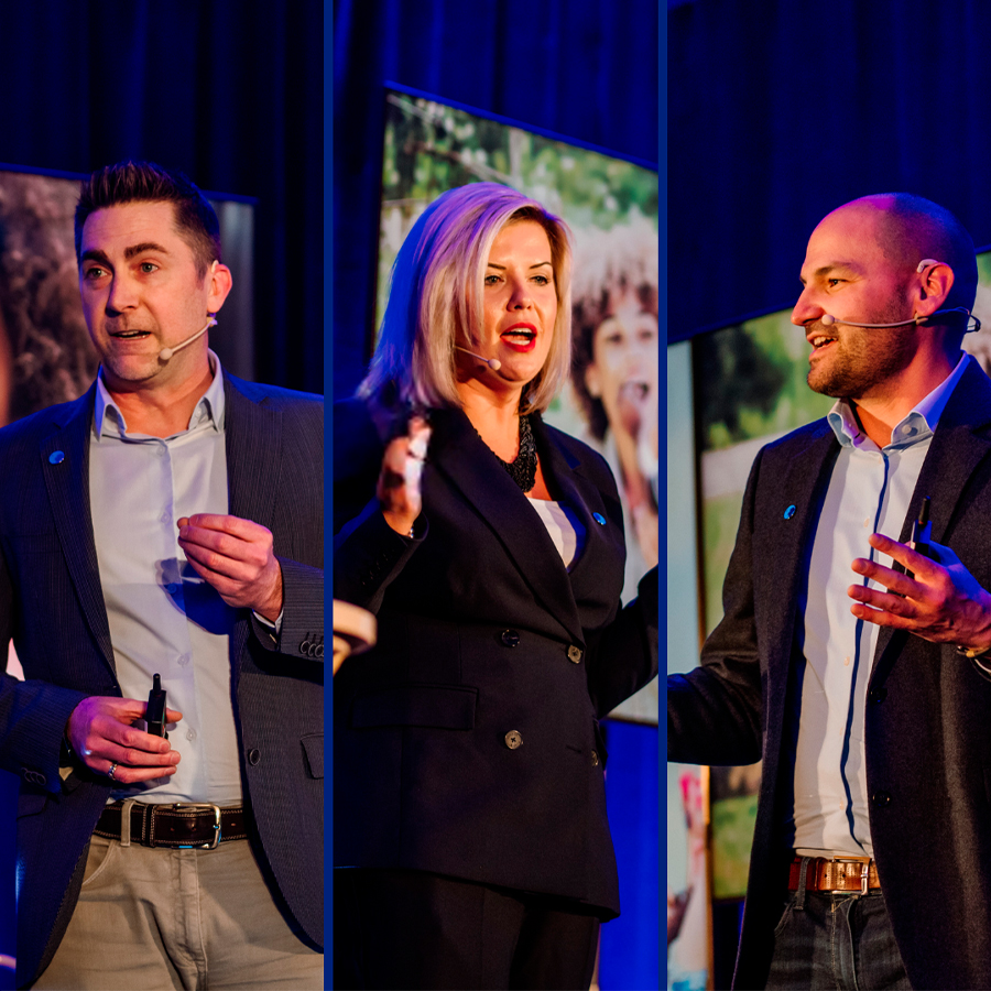 Three Danone Canada leaders stand on stage speaking at an event. From left to right: a man in a black blazer with short, grey straight hair (Frederic Guichard), a woman black blazer and blonde hair (Natalie Holloway), and a man with black blazer and a shaved head (Iannick Melancon). They're all wearing a microphone close to their mouth.