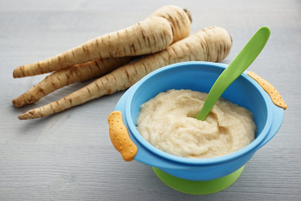 Pastinakenwurzel liegt hinter einer blauen Schüssel mit Pastinakenbrei und grünem Löffel auf grauem Holztisch.