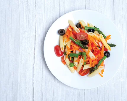 A vibrant plate of pasta salad featuring penne, cherry tomatoes, black olives, asparagus, shredded carrots, and green onions. The dish is served on a white plate placed on a light wooden surface. The colorful ingredients create a fresh and appetizing visual appeal.