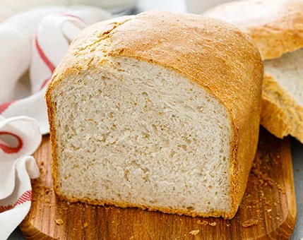 A close-up of a freshly baked loaf of white bread placed on a wooden cutting board. The bread has a golden crust and soft, airy texture visible in the cross-section. A striped kitchen towel is partially visible in the background, adding a homely touch to the setting.