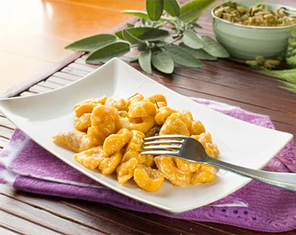 A plate of freshly prepared pasta served with a rich, creamy orange sauce. The dish is presented on a white rectangular plate placed on a purple cloth, with a fork resting on the pasta. Fresh sage leaves and a bowl of seeds are visible in the background, adding a rustic and natural touch to the setting.