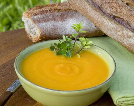 A vibrant bowl of orange soup served in a green ceramic dish, accompanied by rustic bread on a wooden surface. Fresh herbs garnish the soup, adding a touch of greenery. The setting is outdoors, with natural light and a casual dining atmosphere.