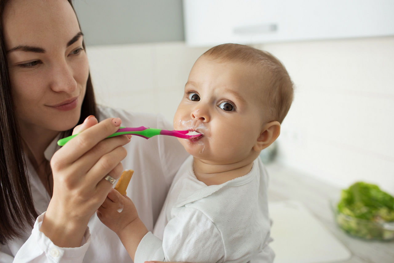 Close up of lovely newborn boy with big brown eyes eating porridge, looking in camera while mother feeding him with spoon and holding kid on hands