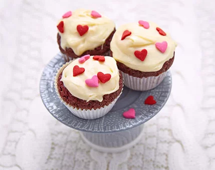Three red velvet cupcakes topped with creamy frosting and decorated with red and pink heart-shaped sprinkles. The cupcakes are displayed on a textured blue cake stand, set against a white knitted background. The vibrant colors and playful design evoke a festive and romantic mood.