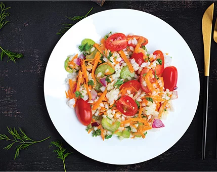 A vibrant vegetable salad featuring cherry tomatoes, shredded carrots, diced cucumbers, and red onions served on a white plate. The dish is placed on a dark textured surface with gold and black cutlery nearby. Fresh dill sprigs are scattered around, adding a touch of greenery to the composition.