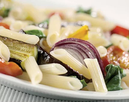 A close-up of penne pasta served with roasted vegetables including zucchini, red onion, and cherry tomatoes. The dish is presented on a white plate, showcasing vibrant colors and fresh ingredients. The image emphasizes a healthy and appetizing meal in a casual dining setting.