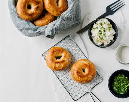 A basket of golden-brown bagels sits alongside a wire rack holding two bagels. The setting includes a bowl of creamy dip garnished with fresh herbs, a small dish of chopped greens, and a fork on a white table. The image evokes a casual dining or breakfast scene with a clean and minimal aesthetic.