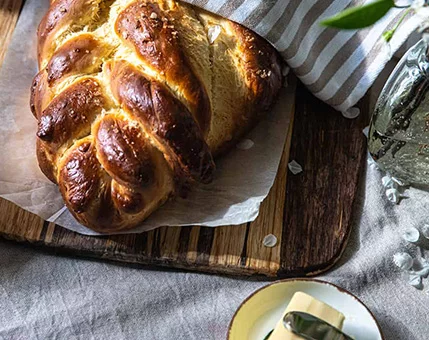 A freshly baked braided bread sits on parchment paper atop a wooden cutting board. The setting features a cozy, rustic table with a striped cloth and a small dish of butter nearby. Warm lighting highlights the golden crust and soft texture of the bread.