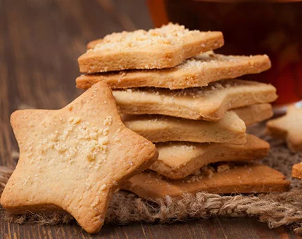 A close-up of golden, star-shaped cookies stacked neatly on a wooden surface. The setting features a rustic aesthetic with burlap fabric and warm tones. Crumbly texture and fine details of the cookies are visible, complemented by a blurred background of a glass containing a dark liquid.