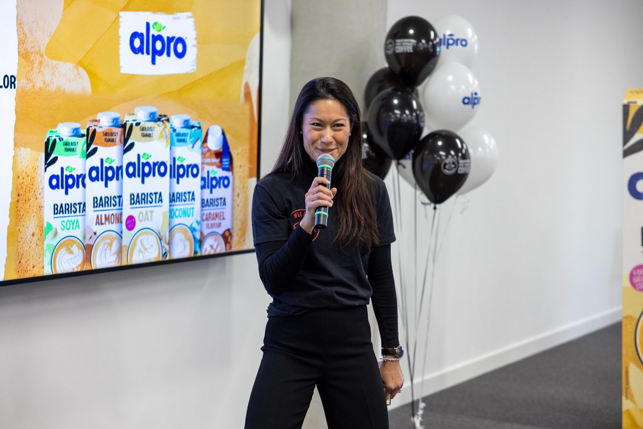 A woman with brown hair stands with a microphone with Alpro branded balloons behind her 