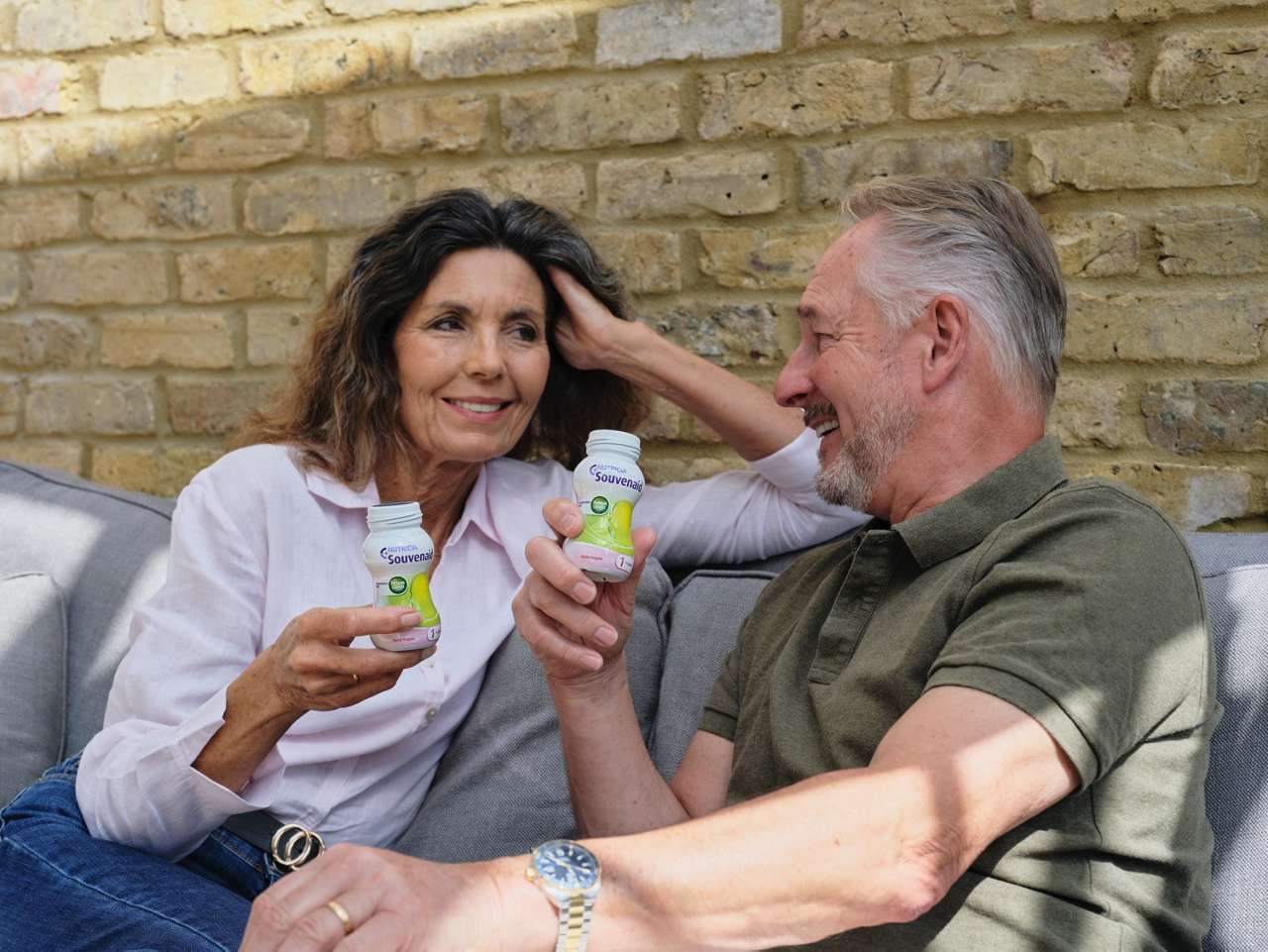 A smiling middle-aged couple sits on a couch against a brick wall, holding yogurt drinks. They look relaxed, enjoying a sunny day together.