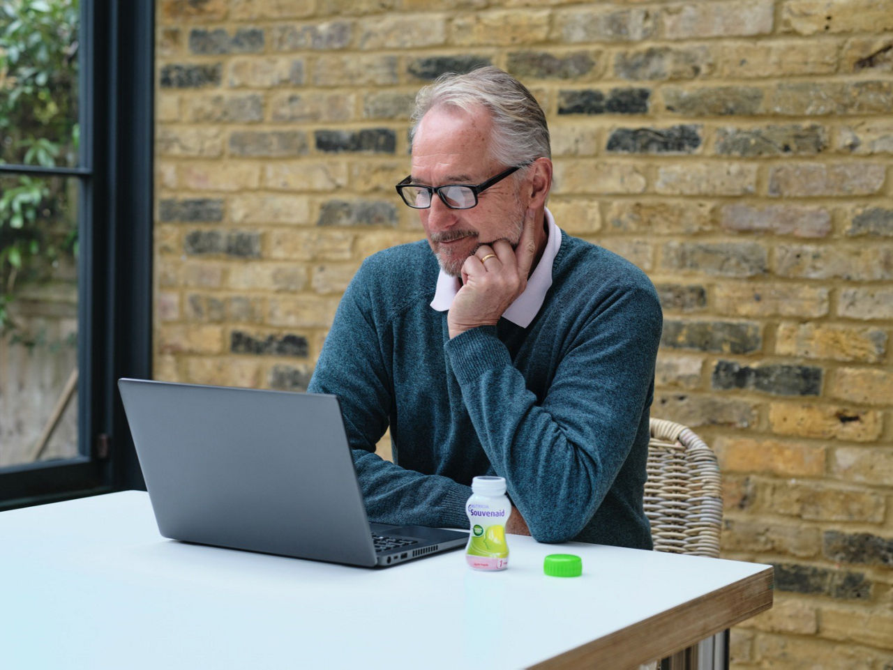 A man sitting at a table and looking at a laptop, with a bottle of Souvenaid® liquid placed next to the computer.