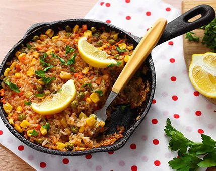 A cast iron skillet filled with a vibrant vegetable dish featuring corn, peas, and rice, garnished with fresh lemon wedges and parsley. The setting includes a polka-dot napkin and wooden cutting board with additional lemon slices and herbs. The dish is presented with a wooden-handled fork resting inside the skillet.