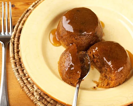 A rich caramel-glazed dessert served on a beige plate with a spoon cutting into one portion. The setting includes a woven placemat and wooden table, evoking a cozy dining atmosphere. The dessert appears moist and indulgent, with warm tones dominating the palette.