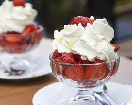 A glass bowl filled with fresh strawberries topped with fluffy whipped cream. The dessert is set outdoors on a wooden table, creating a casual and inviting atmosphere. The vibrant red of the strawberries contrasts beautifully with the white cream, adding visual appeal.