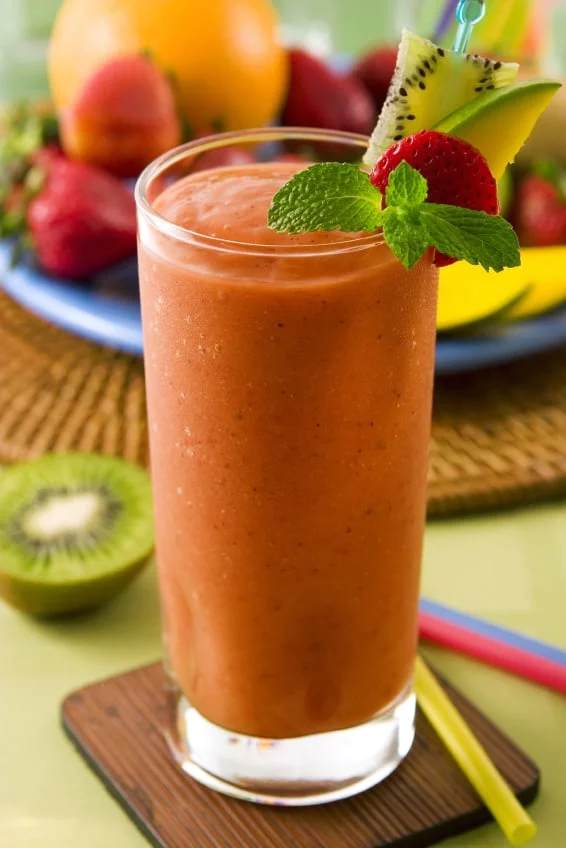 A vibrant fruit smoothie served in a tall glass, garnished with kiwi, strawberry, mango, and mint leaves. The setting includes a colorful arrangement of fresh fruits like oranges and strawberries on a woven mat. Bright and tropical visuals evoke a refreshing and healthy mood.