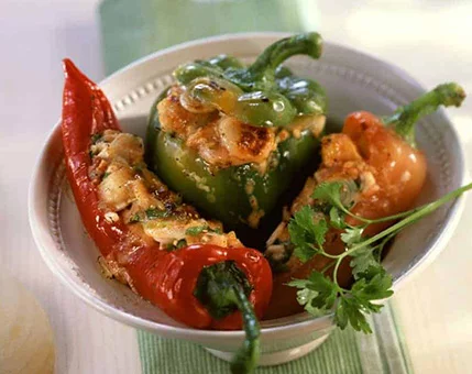 A bowl of vibrant stuffed peppers featuring red, green, and orange varieties filled with a savory mixture. The dish is garnished with fresh parsley and placed on a light green tablecloth. The roasted texture and golden-brown filling add a rustic and appetizing visual appeal.