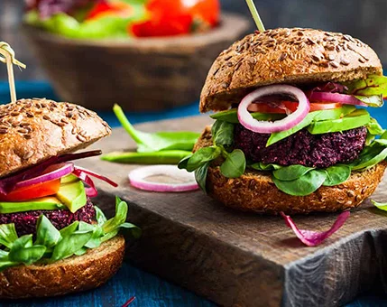 Two vibrant veggie burgers are displayed on a wooden board, featuring beet-based patties, fresh spinach, sliced red onions, and avocado. The setting includes a rustic blue table and blurred vegetables in the background, creating a casual dining atmosphere. The bright colors of the ingredients enhance the visual appeal and suggest a healthy, plant-based meal.