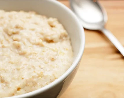 A close-up of creamy oatmeal served in a white bowl, placed on a wooden surface. A silver spoon lies nearby, adding a casual dining element to the composition. The warm tones of the setting complement the texture of the oatmeal.