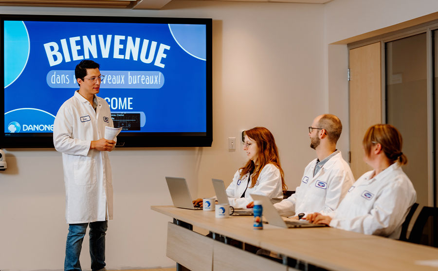 A Danone Canada employee in a lab coat stands by a screen and leads a presentation in a bright room to three colleagues seated at a table with laptops and Danone mugs.