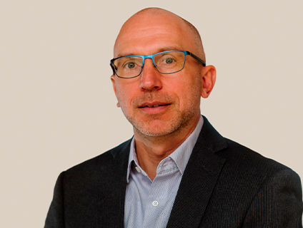 Daniel Crane, Senior Vice President of Operations at Danone Canada, wears glasses and a warm expression in his portrait. He wears a black blazer over a light blue dress shirt and has a shaved head.