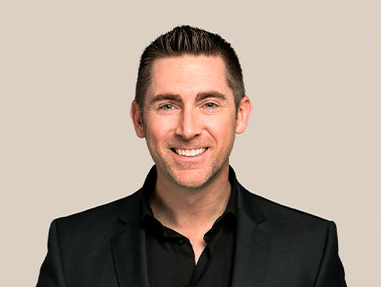 Jeremy Oxley, Senior Vice President of Marketing and E-commerce at Danone Canada, smiles brightly, wearing a black blazer and a black shirt. He has short straight hair, styled with a topknot.