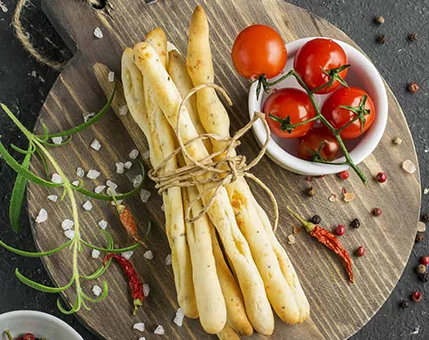A bundle of golden breadsticks tied with twine is placed on a rustic wooden board. Beside them, a small bowl holds fresh cherry tomatoes, surrounded by scattered coarse salt, peppercorns, and dried chili peppers. The setting evokes a Mediterranean-inspired culinary scene with vibrant colors and textures.