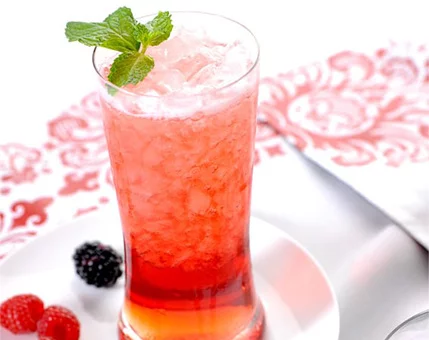A tall glass of iced berry beverage is garnished with fresh mint leaves. The drink features vibrant red and orange hues, complemented by crushed ice. Surrounding the glass are raspberries and a blackberry, placed on a white plate. The setting includes a decorative red and white patterned tablecloth, adding a stylish touch.