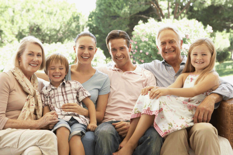 A multigenerational family is seated together on outdoor furniture in a lush garden setting. The group includes adults, children, and seniors, showcasing a warm and relaxed atmosphere. The background features green trees and blooming flowers, enhancing the serene and familial mood.
