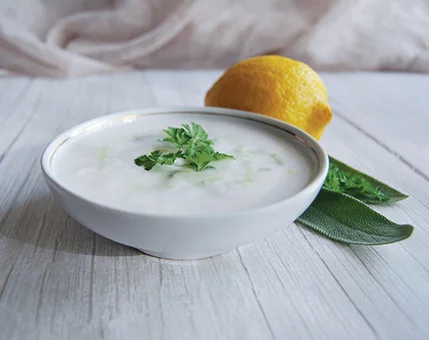 A bowl of creamy yogurt dip garnished with fresh parsley sits on a light wooden surface. A whole lemon and green sage leaves are placed nearby, adding a natural and fresh touch to the composition. The soft background enhances the clean and minimal aesthetic of the image.
