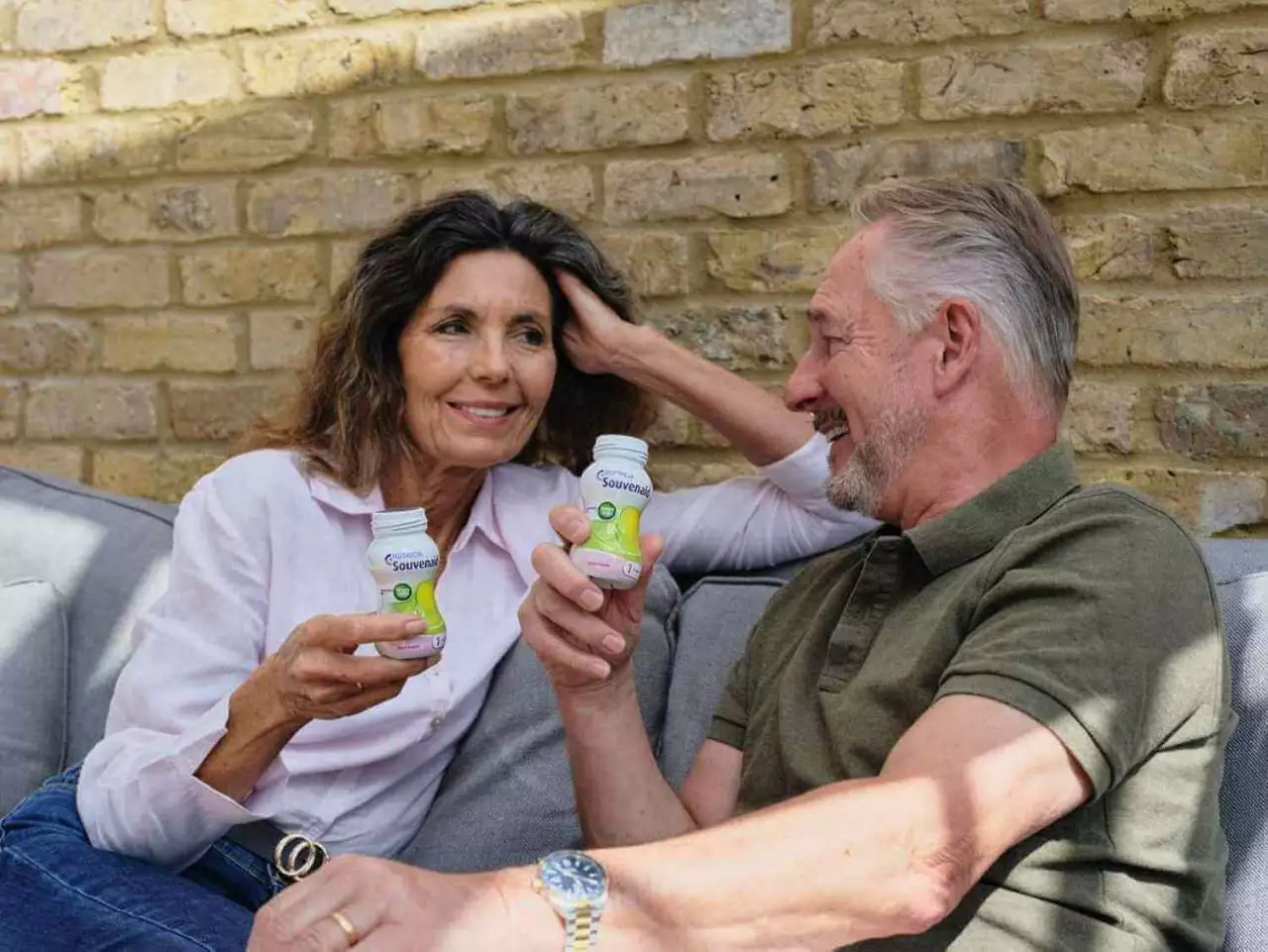 A smiling middle-aged couple sits on a couch against a brick wall, holding yogurt drinks. They look relaxed, enjoying a sunny day together.