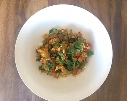 A colorful vegetable rice dish served in a white bowl, placed on a wooden surface. The dish features chopped vegetables, herbs, and grains, creating a vibrant and fresh presentation. The setting suggests a casual dining or home-cooked meal.