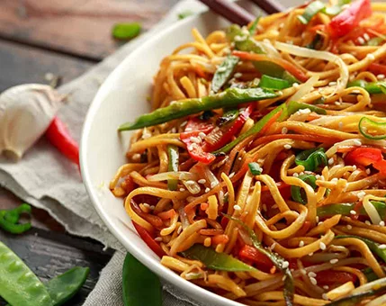 A close-up of a bowl filled with stir-fried noodles, garnished with sesame seeds and colorful vegetables like bell peppers, snap peas, and green onions. The dish is presented on a rustic wooden table with garlic cloves and chili peppers nearby, creating a warm and appetizing setting. The vibrant colors and textures evoke a fresh and flavorful Asian-inspired meal.