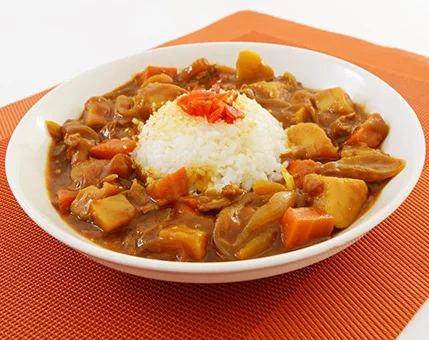 A bowl of Japanese curry served with steamed white rice, garnished with pickled red ginger. The dish features chunks of carrots, potatoes, and onions in a rich, brown curry sauce. Presented on a textured orange placemat, the vibrant colors enhance the warm and comforting mood.