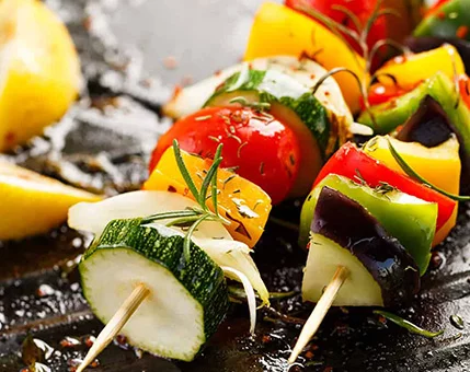 Freshly grilled vegetable skewers featuring zucchini, bell peppers, tomatoes, and onions. The setting showcases a close-up of vibrant, roasted vegetables with visible herbs like rosemary. Lemon wedges are placed in the background, adding a fresh touch to the composition.