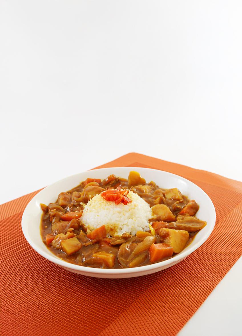 A white bowl filled with curry and rice is placed on an orange textured mat. The curry features chunks of vegetables and a garnish of red chili on top. The setting is minimal with a clean white background, emphasizing the vibrant colors of the dish.