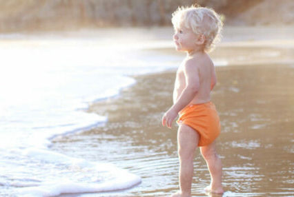 A young child wearing bright orange swimwear stands at the edge of a sandy beach, gazing at the gentle waves. The setting is sunlit, with soft natural lighting and a serene coastal backdrop. The scene evokes a peaceful and playful mood, highlighting the simplicity of childhood moments by the sea.