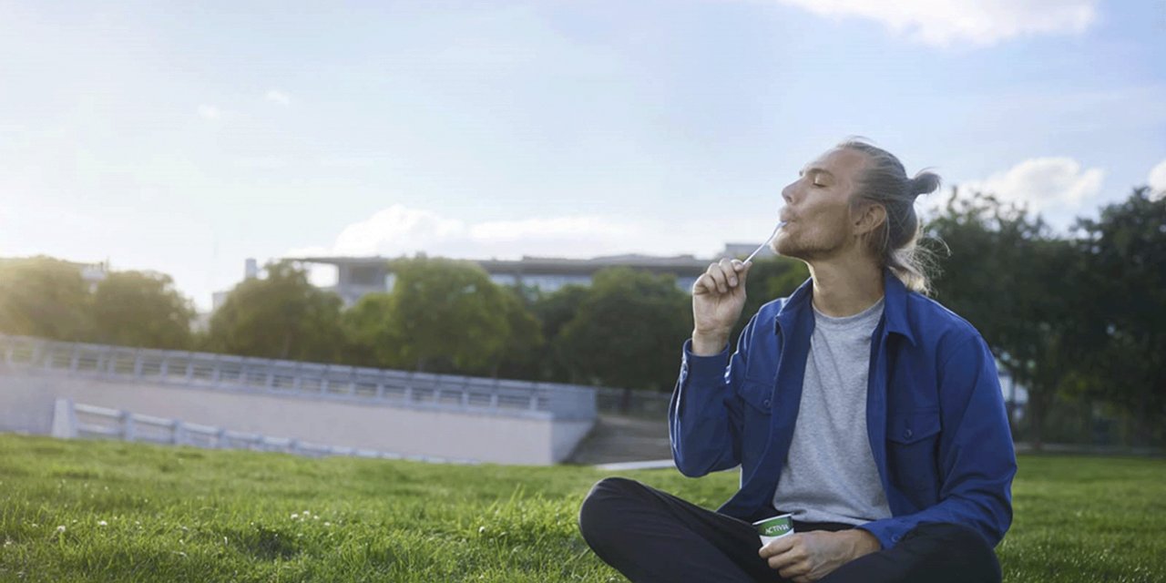 Mann sitzt auf dem Rasen und isst einen Activia Joghurt