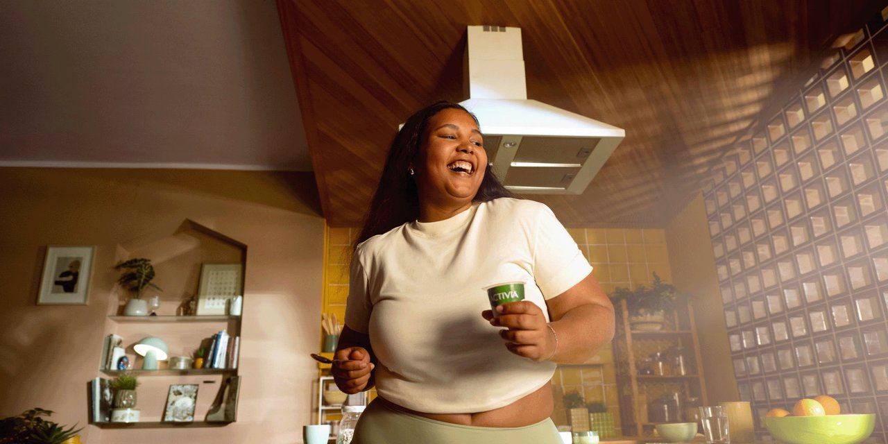 Junge Frau springt im Wohnzimmer mit einem Activia Joghurt in der Hand