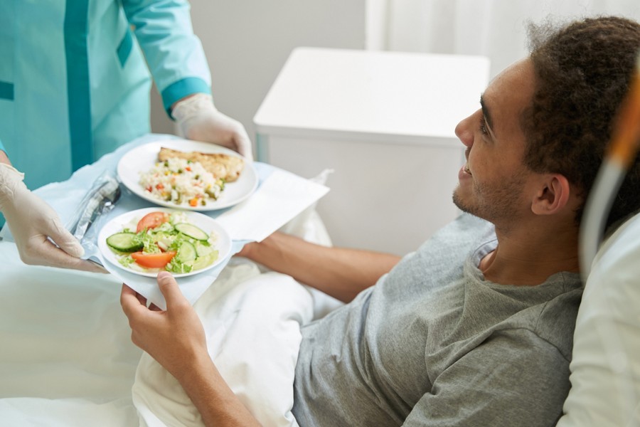 Cheerful male patient in hospital bed taking tray with salad and rice from hands of medic