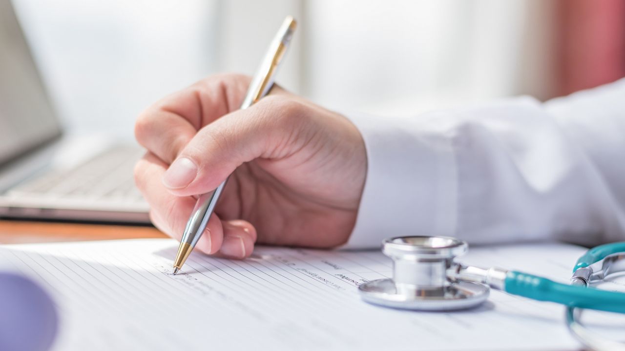 Doctor writing on medical health care record, patients discharge, or prescription form paperwork in hospital clinic office with physician's stethoscope on desk